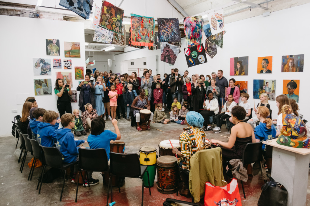 Audience gathered around a drumming circle in a gallery hung with colourful paintings