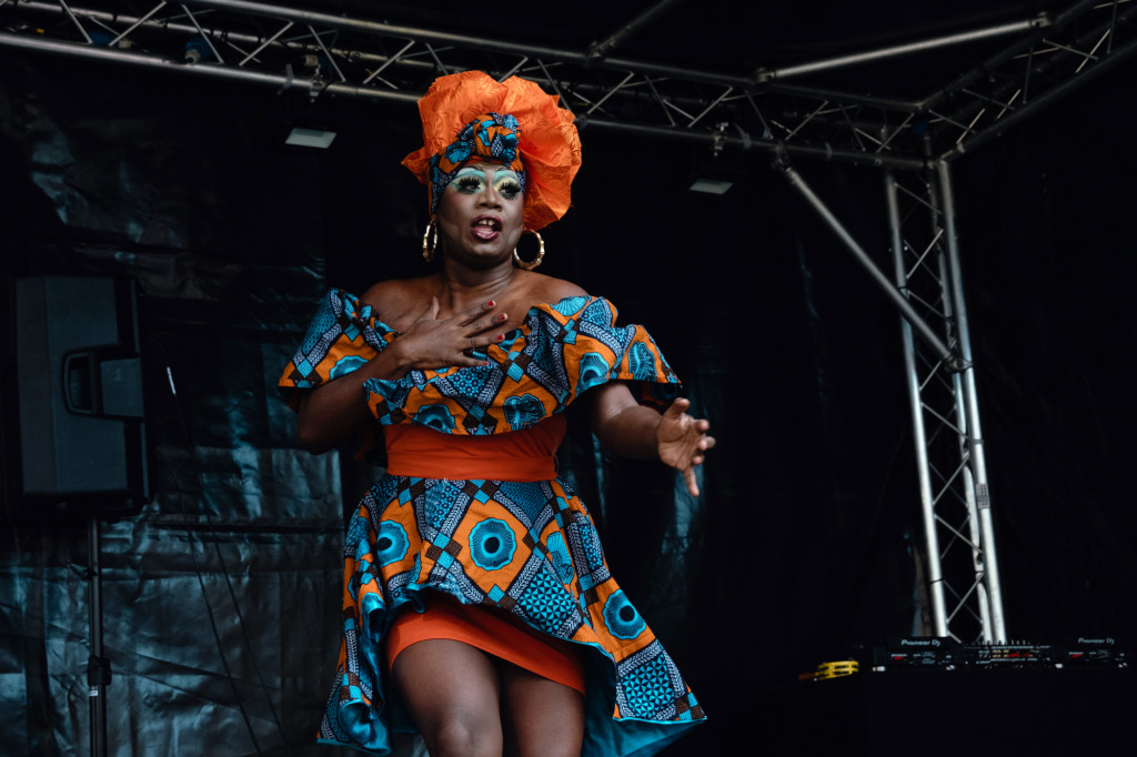 Performer in a patterned dress and orange headwrap singing on stage